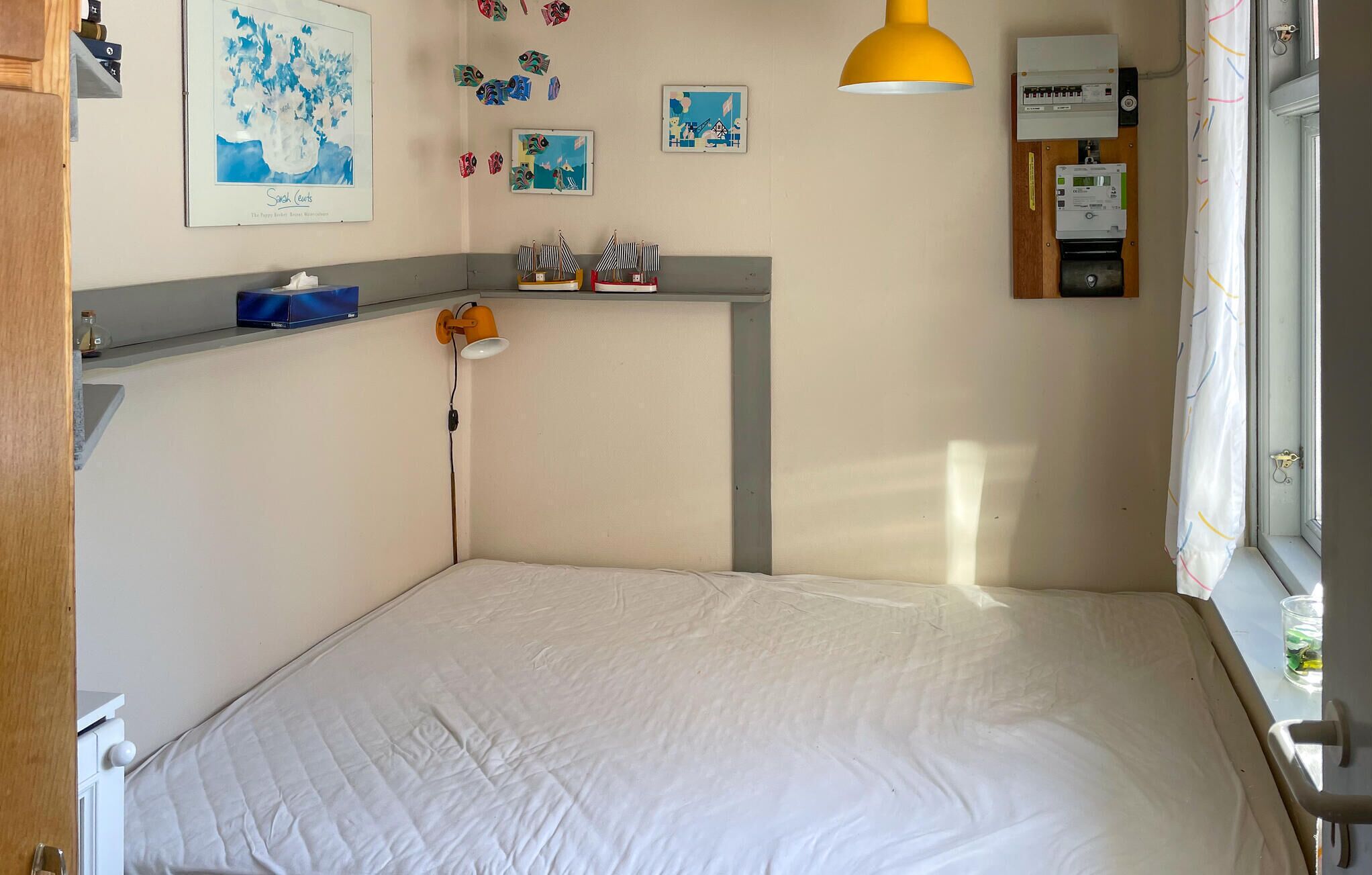 Un dormitorio pequeño y ordenado con una cama, una repisa con objetos personales, una lámpara de pared y una ventana con cortinas.