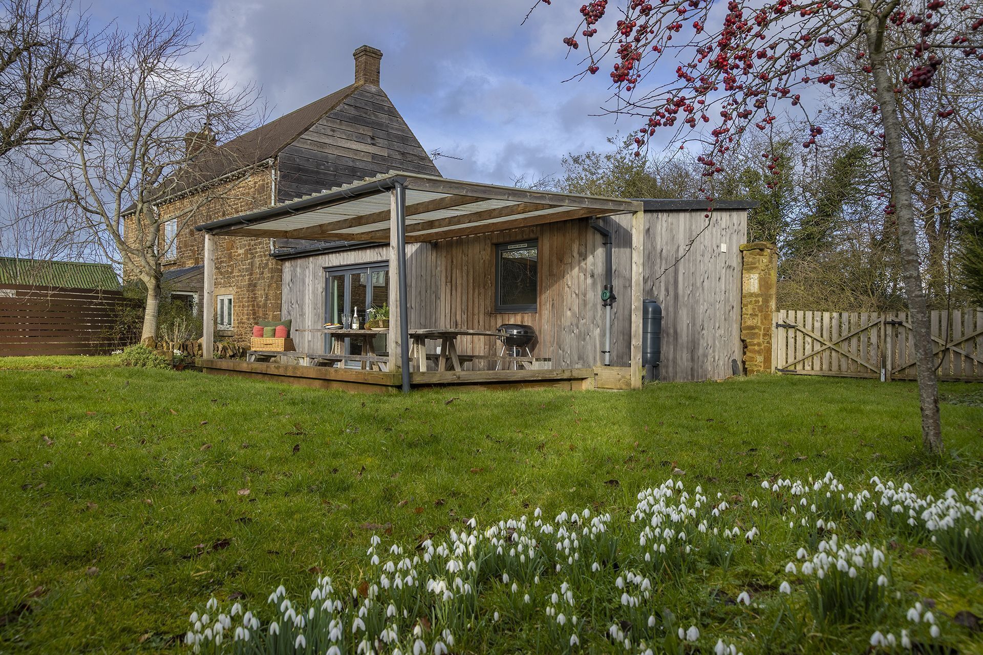 A rustic wooden house with a covered patio, a stone chimney, and a fenced yard.