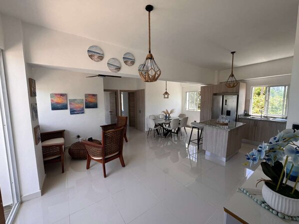A spacious kitchen with a dining area, white tiled floors, and a large window providing natural light.