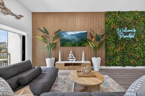 Ein modernes Wohnzimmer mit einem großen Flachbildfernseher, einer Couch, einem Couchtisch und einer Wand mit dekorativen Elementen, auf der ein Schild mit der Aufschrift 'Welcome to Paradise' angebracht ist.