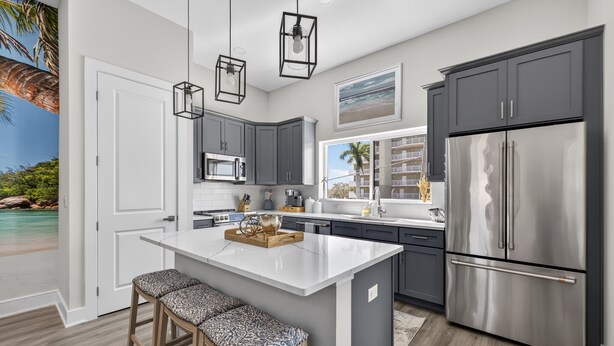 Une cuisine moderne dotée d’un îlot central, de meubles gris, d’appareils électroménagers en acier inoxydable et offrant une vue sur la plage grâce à une grande fenêtre.