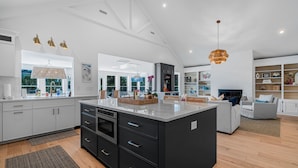 A modern kitchen with a large island, white cabinetry, and a dining area with a fireplace. - Modern Coastal Beach Home! Across from Beach Access w/Ocean Views & a Huge Yard w/Private Pool! (Isle Of Palms)