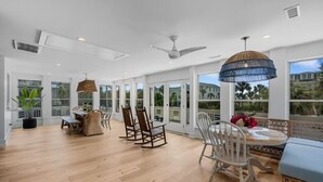 A spacious room with a wooden floor, a dining area with chairs and a table, a ceiling fan, and large windows. - Modern Coastal Beach Home! Across from Beach Access w/Ocean Views & a Huge Yard w/Private Pool! (Isle Of Palms)