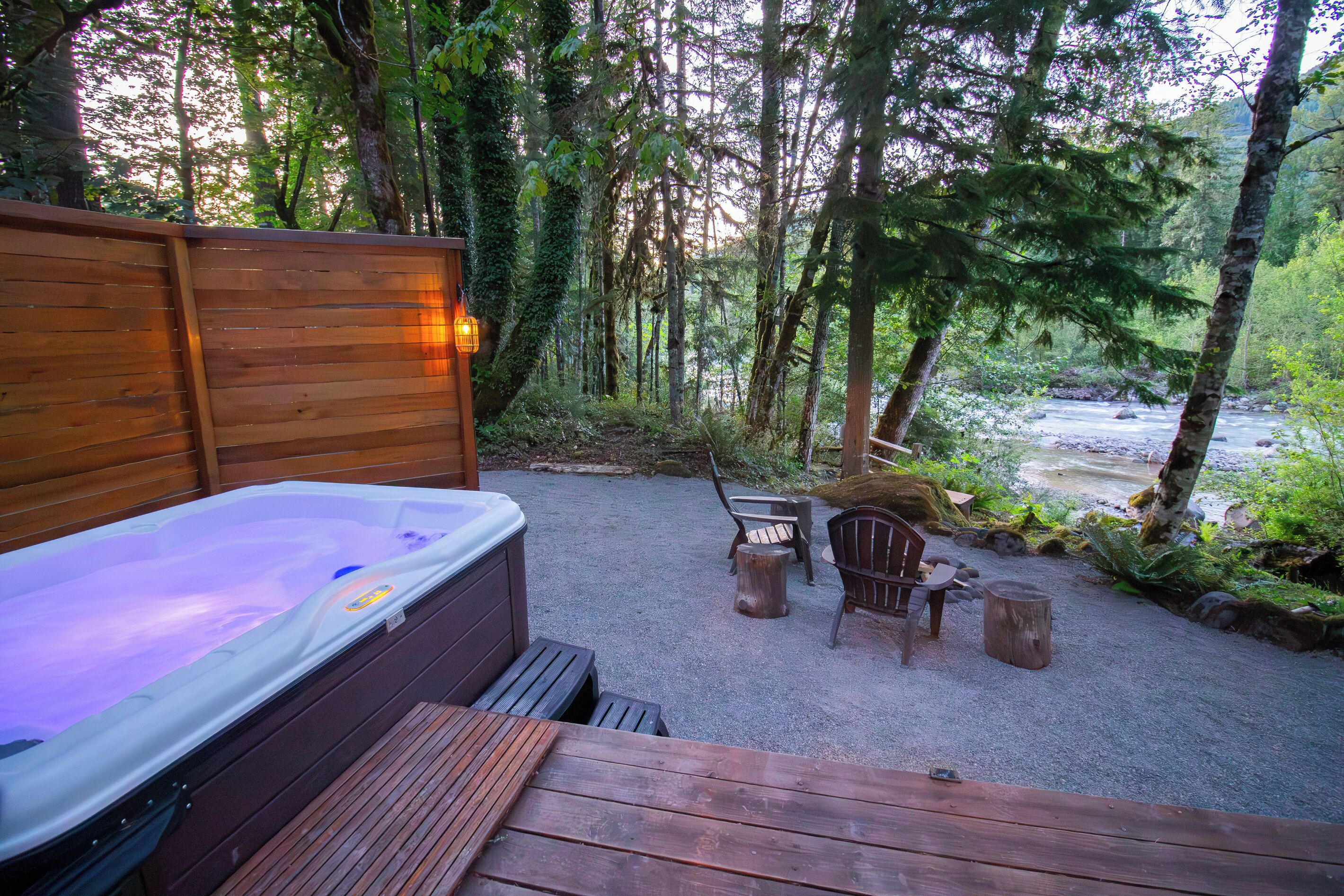A hot tub with a wooden deck, surrounded by a forested area with a river view.