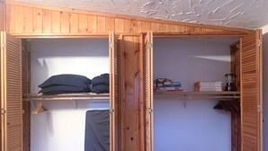 Una habitación con un armario de madera, una cama con una manta gris y una estantería con libros y una botella.