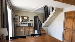 Ein Essbereich mit Holztisch und -stühlen, eine Treppe mit schwarzen Geländern, eine Holztür und ein Küchenbereich mit Regalen und einem Spiegel.