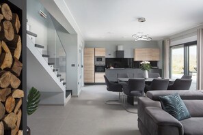 Eine moderne Küche mit einer zentralen Kochinsel, ein Essbereich mit grau gepolsterten Stühlen und eine Treppe mit Glasgeländern.