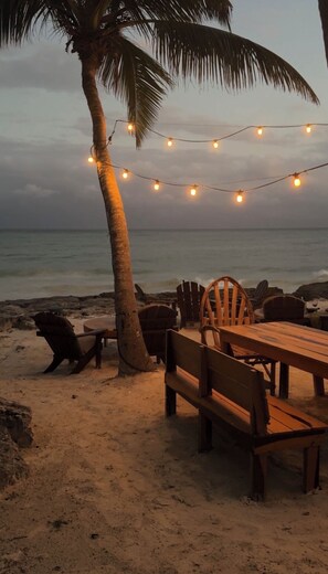 Ein Essbereich am Strand mit Holztischen und -stühlen, Lichterketten und einer Palme.