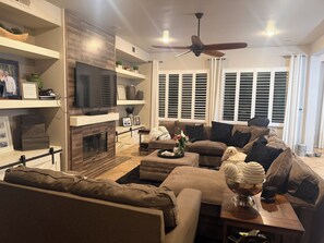 A modern living room with a fireplace, a large flat-screen TV, and a ceiling fan. - Charming N. Indio 5-bedroom Beauty (Indio)