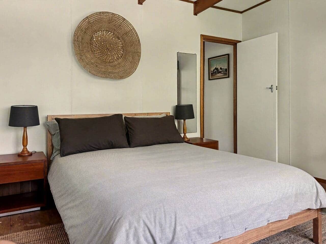 A bedroom with a bed, two bedside tables with lamps, a woven wall hanging, a door with a picture, and a framed artwork on the wall.