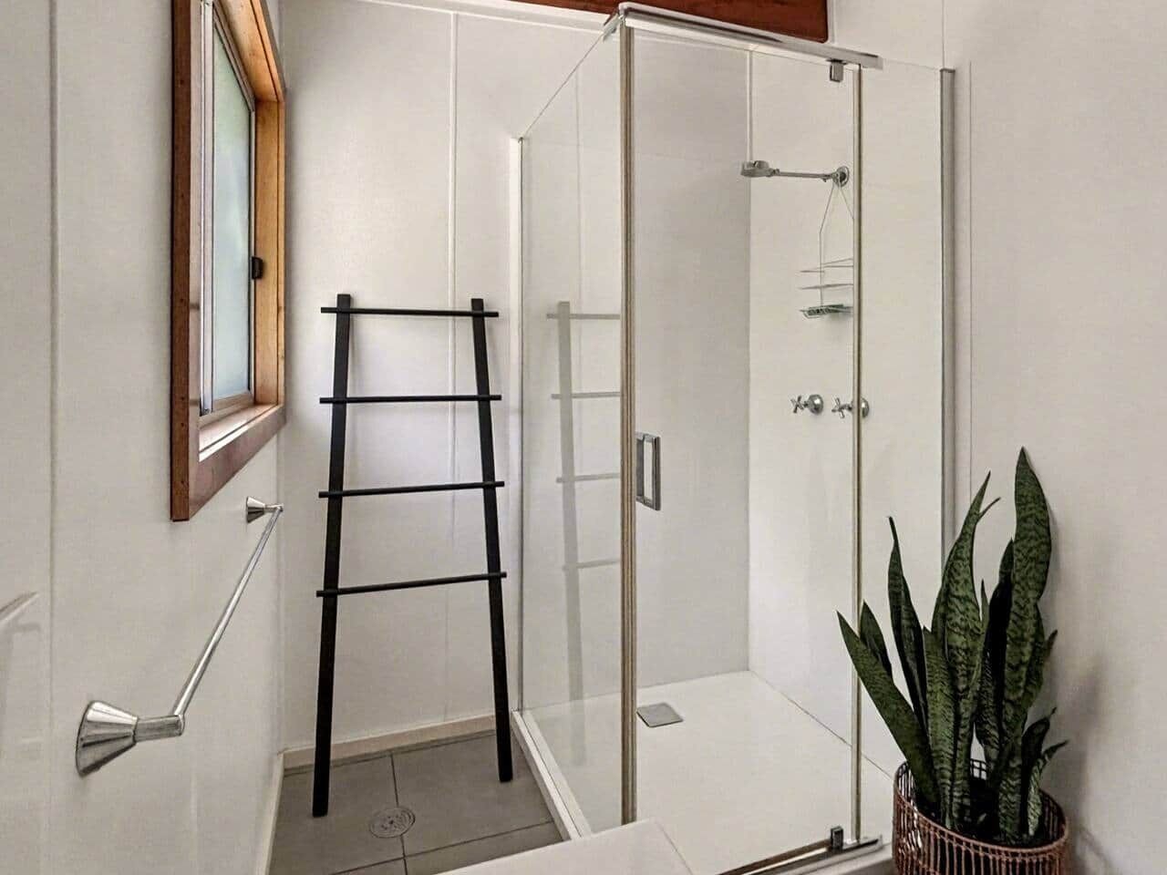 A modern bathroom with a glass shower enclosure, a wooden-framed window, a metal towel rack, and a potted plant.