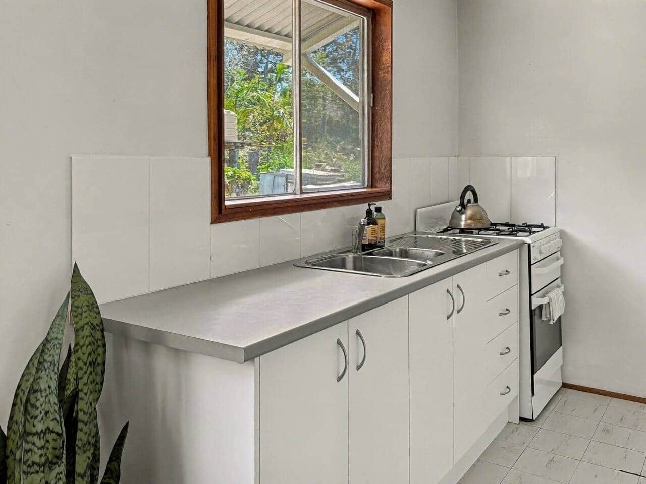 Une cuisine avec des meubles blancs, un plan de travail en acier inoxydable, une cuisinière à gaz et une fenêtre donnant sur un paysage verdoyant.