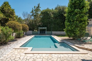 Ein rechteckiger Swimmingpool mit Steinbordur, umgeben von Kies, einer überdachten Sitzgelegenheit sowie verschiedenen Bäumen und Sträuchern.