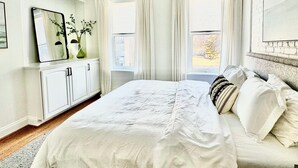 Une chambre à coucher comprenant un lit, un ventilateur de plafond, une fenêtre avec des rideaux, une commode et un miroir.