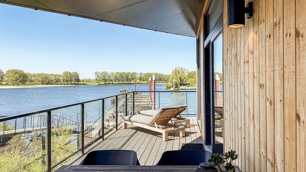 Une terrasse en bois avec une balustrade en verre donnant sur un lac, une chaise longue et une table.