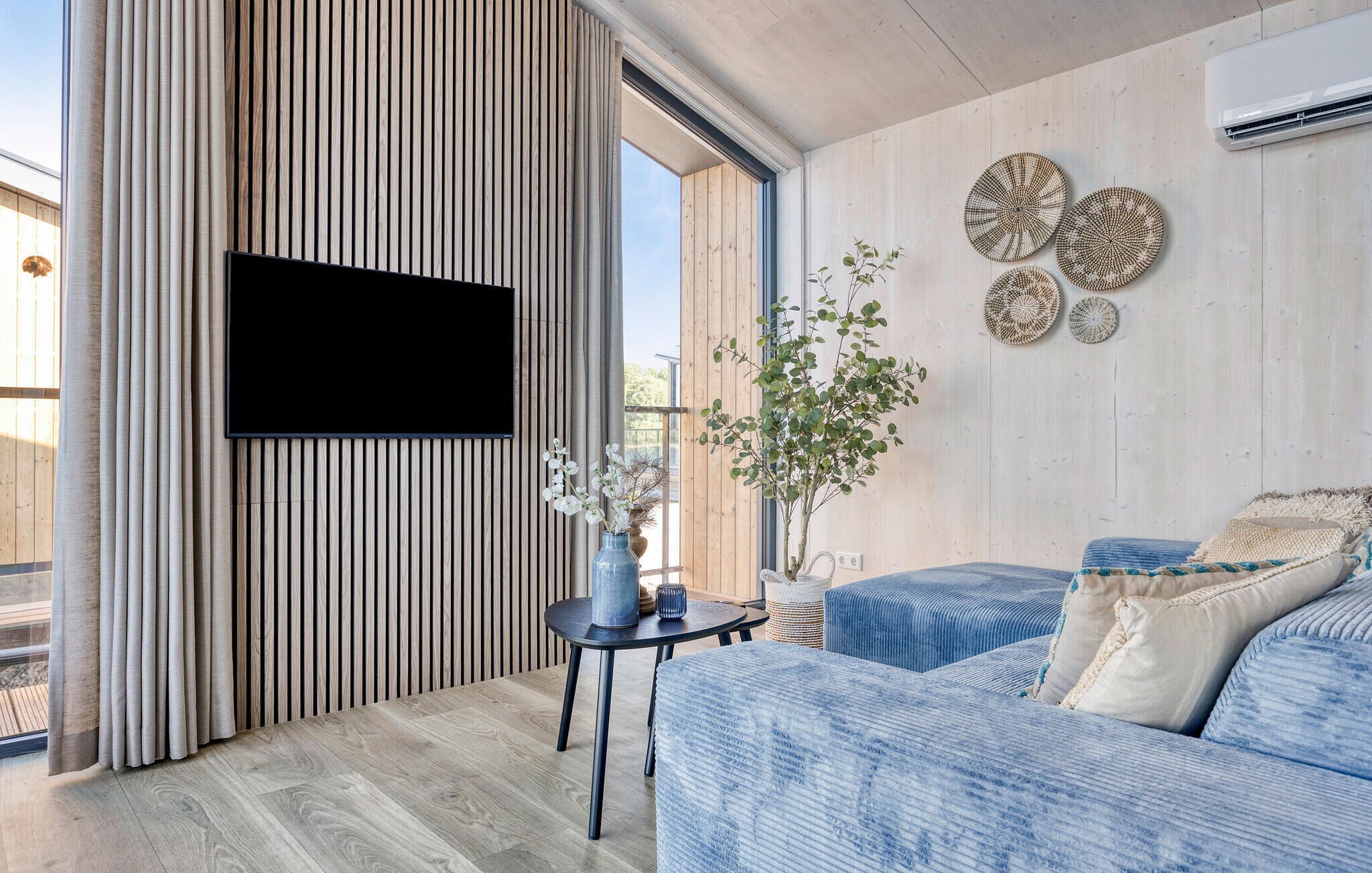A modern living room with a large flat-screen TV mounted on a wooden wall, a blue sofa, and a small side table with a vase of flowers.