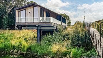 Een moderne, verhoogde cabin met balkon, gelegen boven water, omringd door weelderige begroeiing.