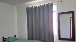 Une petite chambre avec un lit simple, une tête de lit en bois, une fenêtre avec des rideaux gris, un miroir au mur et un sol carrelé.