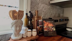 Eine Küchenarbeitsplatte mit Schneidebrett aus Holz, einer weißen Keramikfigur, einer Olivenölflasche, einer Vase mit Trauben, einem Kochbuch mit dem Titel „Around the Fire“ und einem modernen Herd.