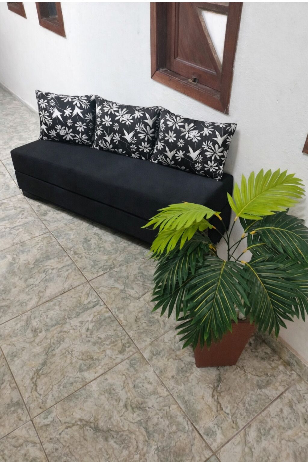 A black sofa with floral-patterned cushions, a potted plant with large green leaves, and a tiled floor.
