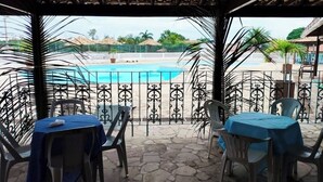 A covered outdoor seating area with tables and chairs, overlooking a swimming pool. - CostaeCunhaIpe104  (Iguaba Grande)