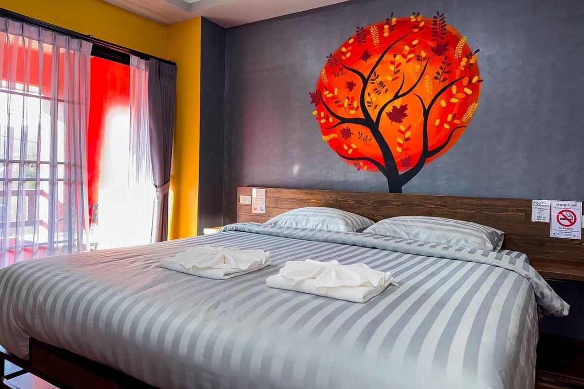 A hotel room with a bed, striped bedding, a large painting of a tree, and a window with curtains.