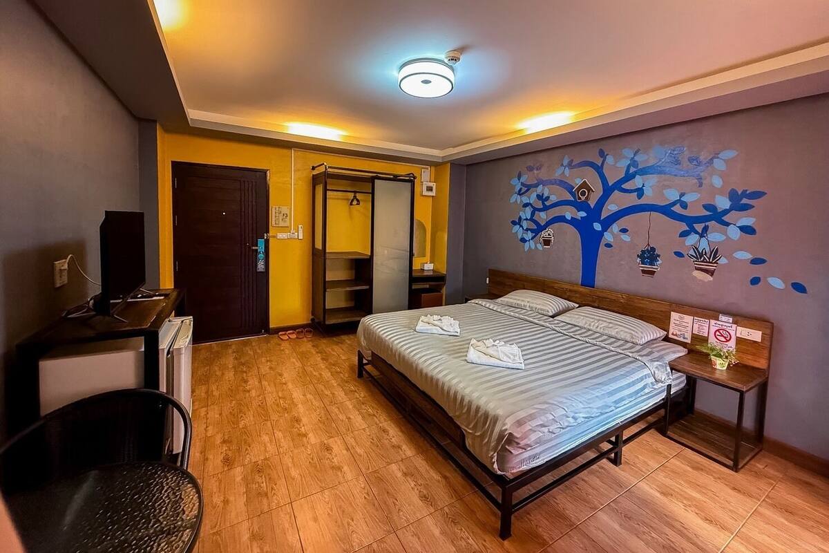 A bedroom with a bed, a desk, a chair, a wardrobe, and a wall with a tree mural.