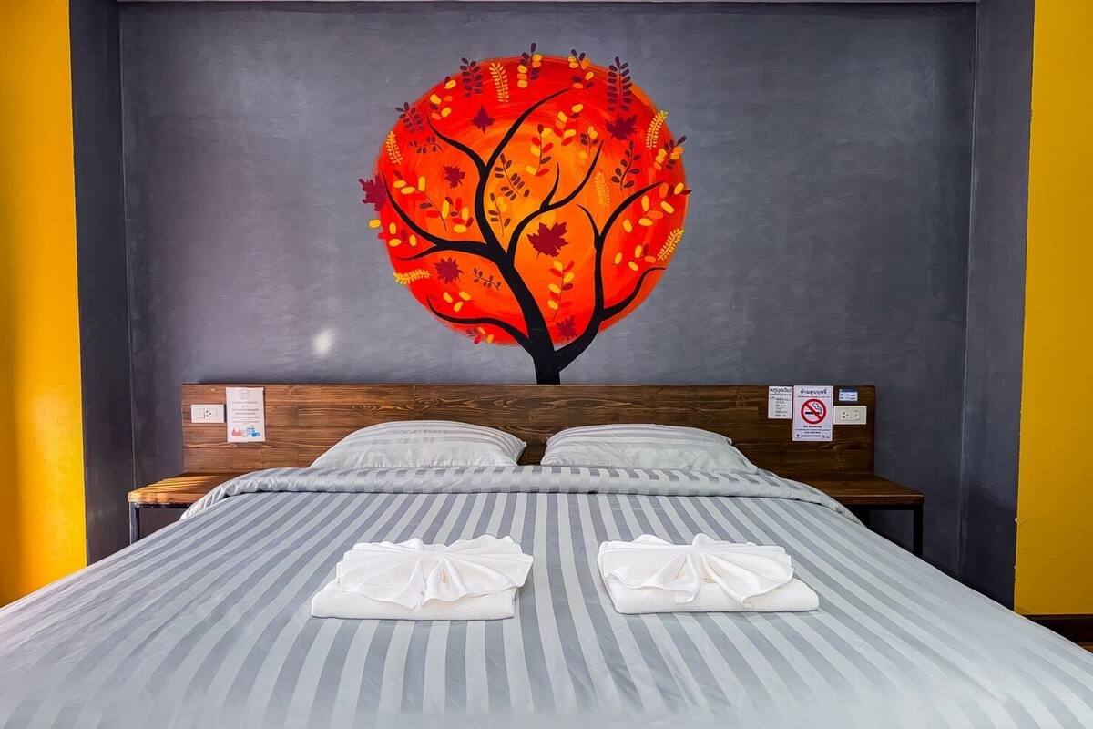 A hotel room with a bed, two pillows, a headboard with a sign, and a wall painting of a tree.