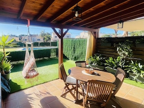 Eine überdachte Terrasse mit einem Holztisch und Stühlen, umgeben von Topfpflanzen und einem Hängesessel.