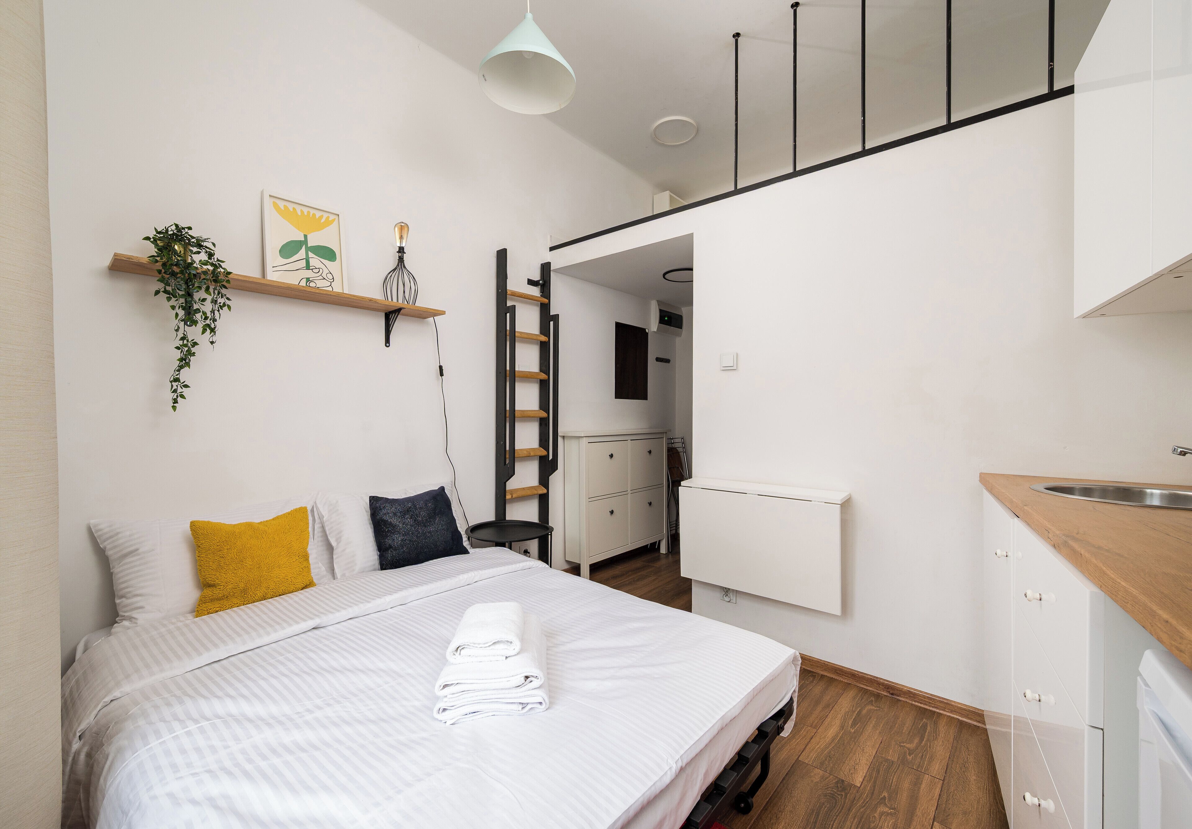 Ein modernes Schlafzimmer mit einem Bett, einem Holzregal mit Pflanzen, einer Leiter und Blick in die Küche.