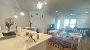 A modern kitchen with a white countertop, a dining area with a table and chairs, and a living room with a sofa and a TV.