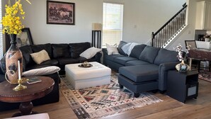 A living room with a dark-colored sofa set, a white ottoman, a patterned rug, and a vase with yellow flowers.