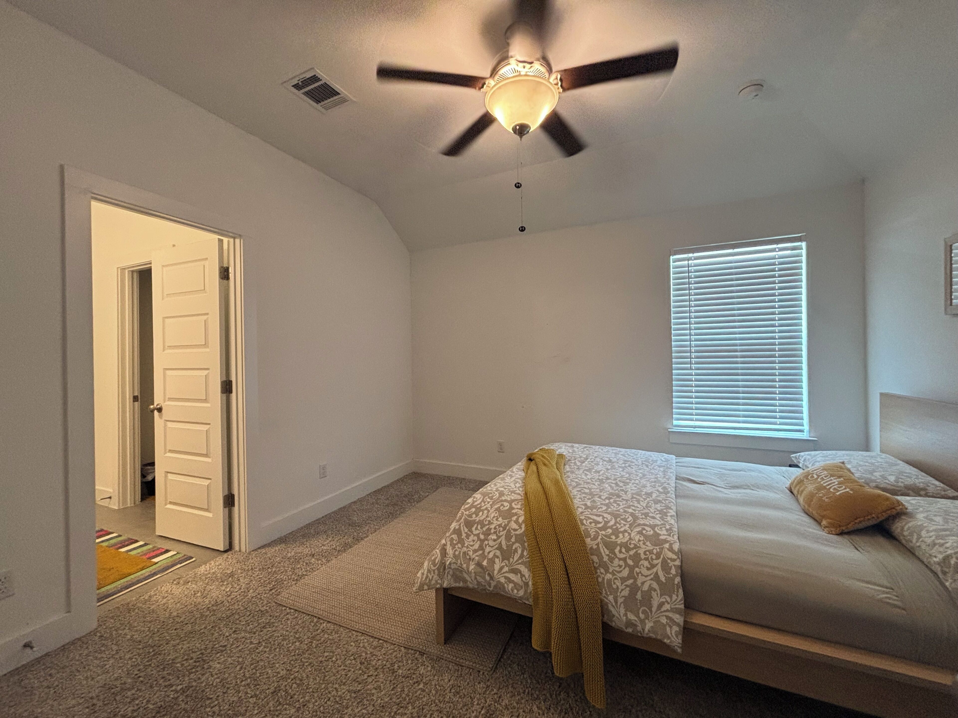 A bedroom with a bed, a ceiling fan, a window with blinds, and a door leading to another room.