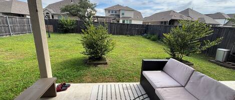 A backyard patio with a cushioned outdoor seating area, a small tree, and a wooden fence.