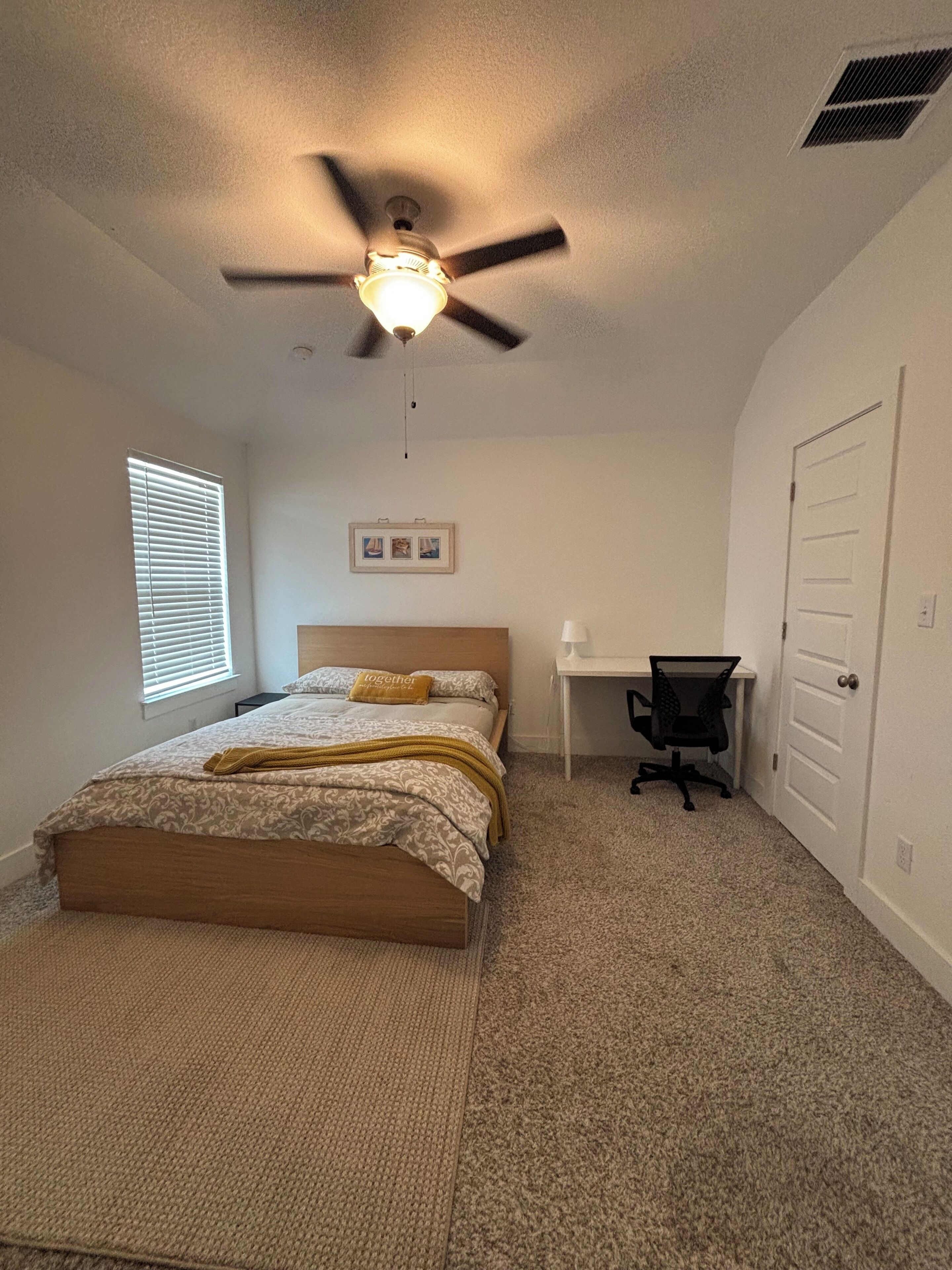 Ein Schlafzimmer mit Bett, Schreibtisch, Stuhl, Deckenventilator und einer Tür.