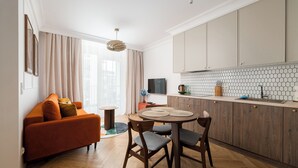 Ein modernes Wohnzimmer mit einer Couch, einem Esstisch mit Stühlen, einer Küchenzeile mit Schränken und einer gefliesten Spritzschutzwand sowie einem großen Fenster mit Vorhängen.