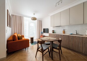 A modern living room with a sofa, a dining table with chairs, a kitchen area with cabinets and a tiled backsplash, and a large window with curtains. - Warszawa Rakowiec Superior Apartment (Warszawa)