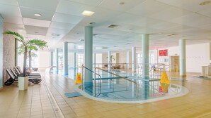 Amplia zona de piscina interior con agua cristalina, rodeada de suelos de baldosas y columnas, y equipada con barandales de seguridad.