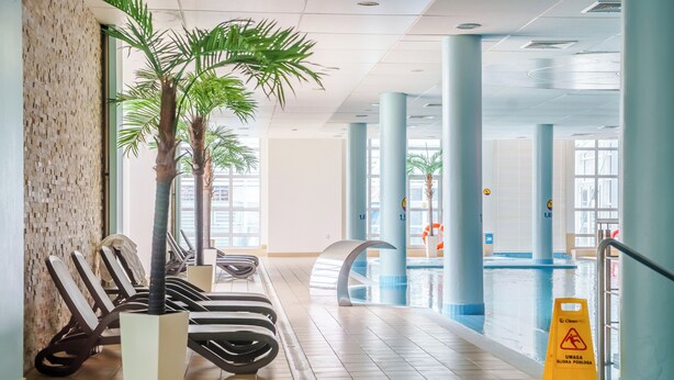 Un área de piscina interior con sillones de descanso, palmeras y un letrero de precaución.
