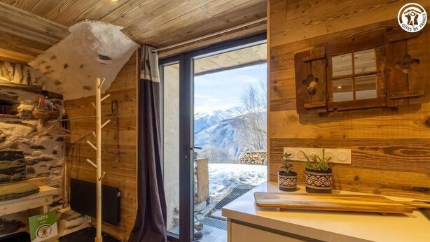 A cozy cabin interior with a stone wall, wooden ceiling, a wooden coat rack, a television, a window with a view of mountains, and a kitchen counter with potted plants. - LE GITE DE LA MONTAGNETTE (BEAUNE)