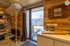 A cozy cabin interior with a stone wall, wooden ceiling, a wooden coat rack, a television, a window with a view of mountains, and a kitchen counter with potted plants. - LE GITE DE LA MONTAGNETTE (BEAUNE)
