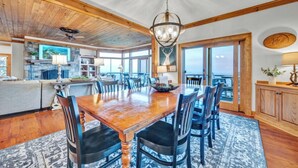 A spacious dining area with a wooden table and chairs, a chandelier, and a view of the ocean through large windows. - 3BR Home w/180° Mountain Views | Hot Tub, Poker Room, Sauna | 5 Min to Ski Resort (Beech Mountain)