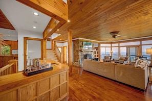 A spacious living area with a wooden ceiling, a stone fireplace, and a large window overlooking a scenic view. - 3BR Home w/180° Mountain Views | Hot Tub, Poker Room, Sauna | 5 Min to Ski Resort (Beech Mountain)