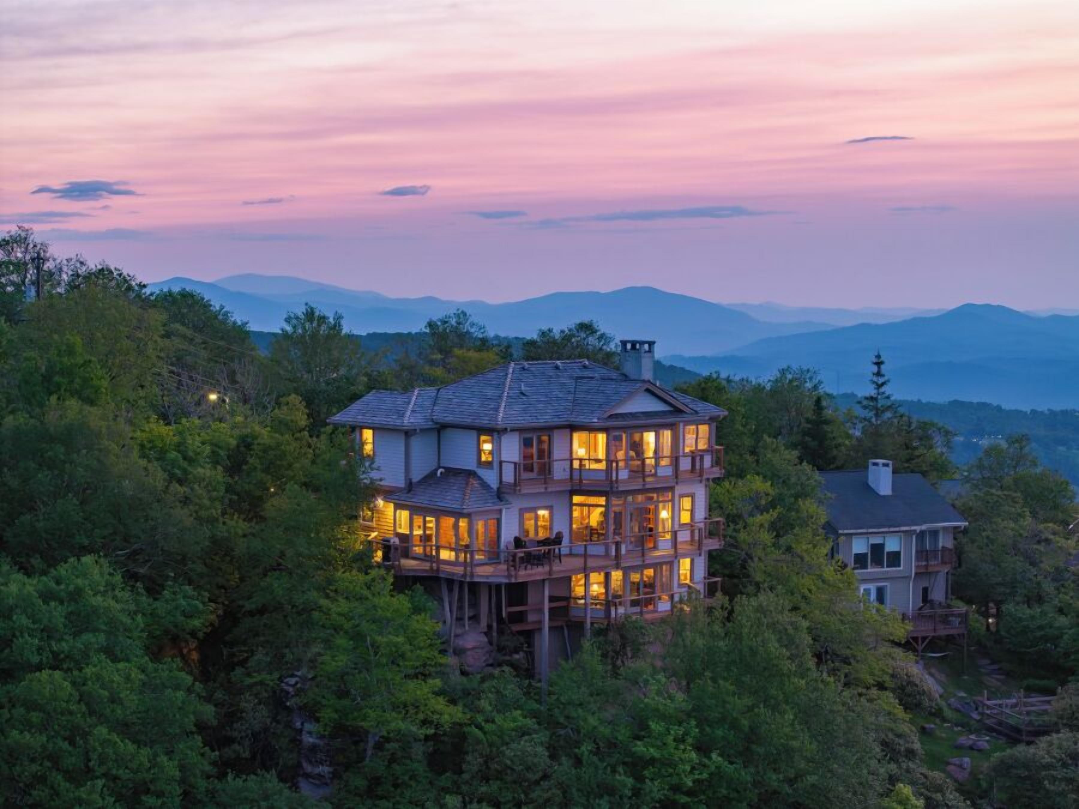 A multi-story house with a deck, surrounded by dense forest and mountains in the background.