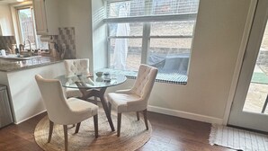 En matplats med ett glasbord och fyra stolar, ett kök i bakgrunden och ett stort fönster med utsikt över omgivningen.