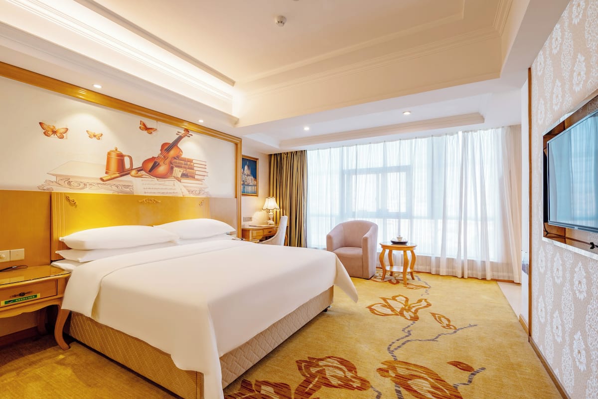 Een hotelkamer met een groot bed, een televisie, een bureau met lamp, een stoel en een raam met gordijnen.