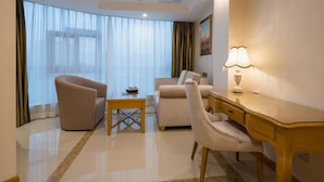 Una habitación de hotel con sofá, sillón, escritorio y un ventanal con cortinas translúcidas.