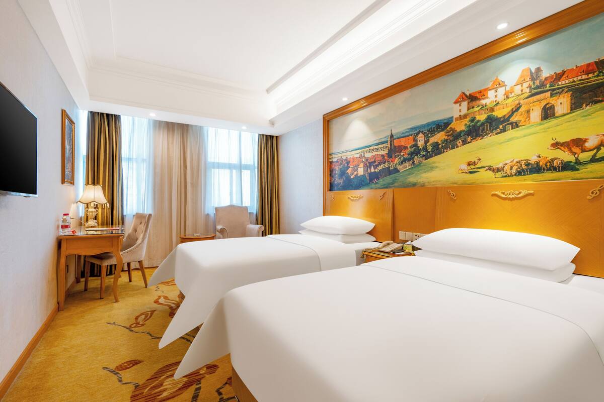 Een hotelkamer met twee bedden, een bureau met stoel, een televisie en een schilderij aan de muur.