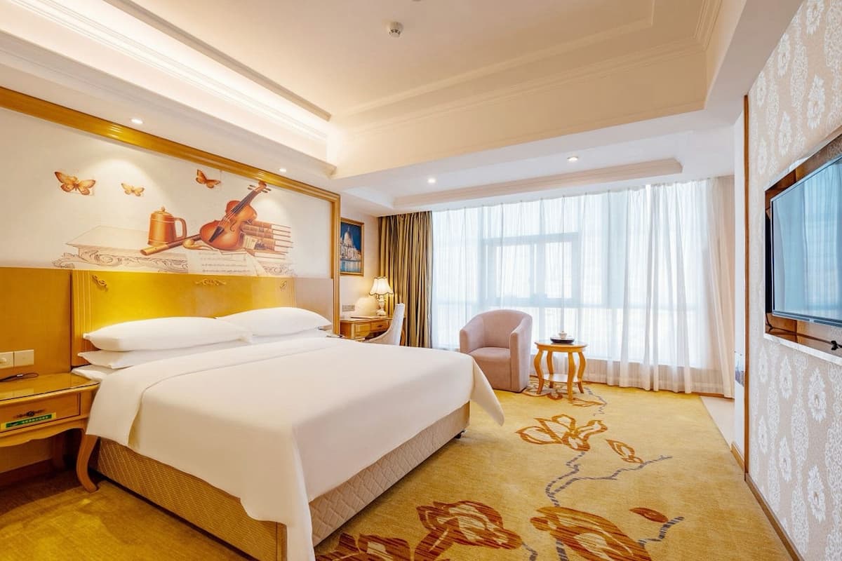 Een hotelkamer met een groot bed, een televisie, een bureau met lamp, een stoel en een raam met gordijnen.