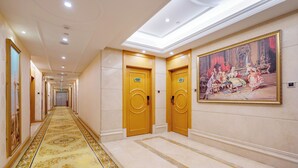 Un pasillo de hotel con una alfombra larga y ornamentada, dos puertas de madera con letreros y un cuadro clásico grande en la pared.